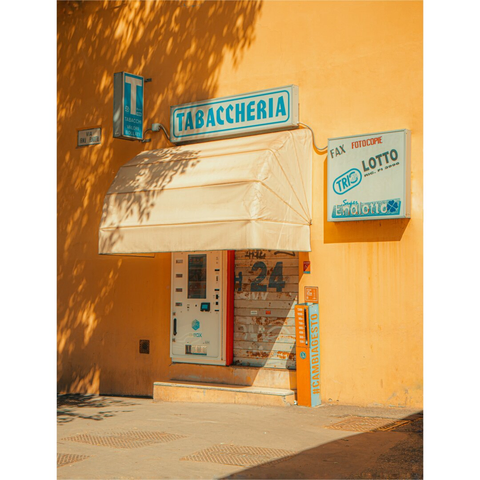 A photograph of a small, sunlit tabacheria (tobacco shop) with orangey/yellow walls. The shop has a beige awning a serrated door covering and a dispenser machine on its left.. Above the door, a blue sign reads "TABACCHERIA." To the right, a white sign with blue and red text reads "ENALOTTO." Shadows from nearby trees cast patterns on the wall on the left. The pavement in front is grey.