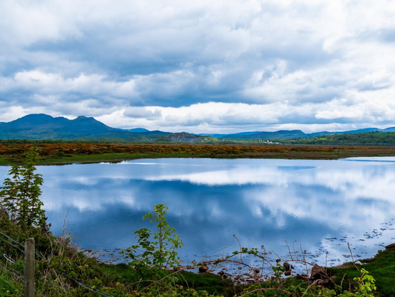 A scenic view of the Glaslyn Estuary in Porthmadog today. The calm, blue water mirrors large white and grey clouds, framed by lush green banks and the silhouetted peaks of the Eryri (Snowdonia) mountain range.
