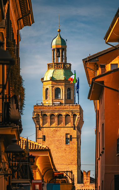 This is a photograph a tower (La Torre dell'Orologio) between warm coloured buildings on either side. (Via D'Azeglio). The.tower has arched windows and a greenish patina dome. An Italian flag and a European Union flag are flying prominently from the side of the tower. There's a small bell which sits within the open-air belfry at the very top of the it. The surrounding buildings are in shades of ochre/yellow and orange. Everything is illuminated by golden light.