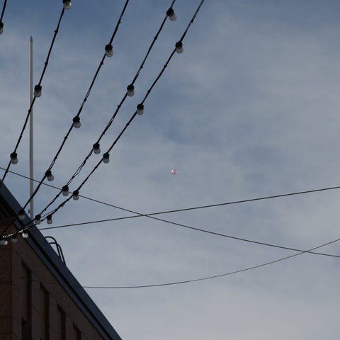 Crisscrossing string lights stretch across a blue sky, anchored to a building on the left. Behind them, in the middle, a pink helium balloon drifts up to the sky.