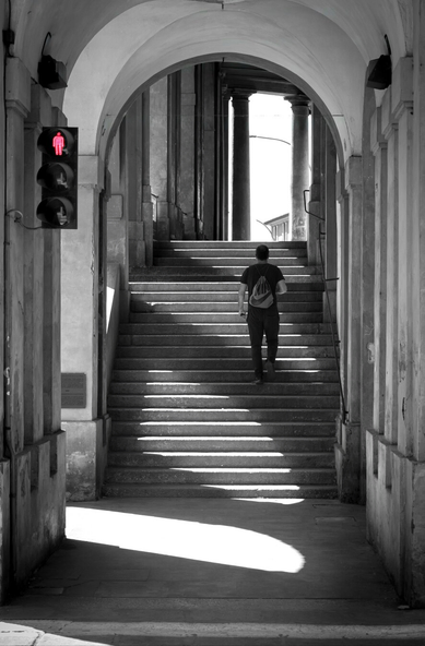 This is a black-and-white photograph of a person walking up a wide set of stone stairs underneath annarchway (This is in Meloncello) The person is seen from behind - they are wearing a dark t-shirt, dark trousers, and a small backpack. On the left side of the photo there is a pedestrian traffic light and it has the glowing red "stop" icon
 It is the only splash of colour in the image. Sunlight is coming in from the left of the photo and casts sharp shadows across the stairs and on the ground.