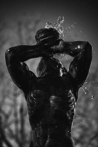 A high contrast monochrome photograph of a fountain statue of the Greek god Triton. The figure is holding up a cup to its head and from the cup flows water, spraying all over in the windy conditions. The statue is photographed from the front and from the torso and up. The statue is rendered quite dark creating a contrasty image in the harsh sunlight and fills a large part of the image.

The short shutter speed freezes the spraying water and the large aperture renders the background with trees and sky quit blurry.