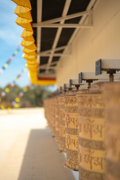 I took this photo at the Great Stupa of Universal Compassion near Bendigo, in Victoria Australia. you can see a long line of Tibetan prayer wheels along one of the white weels of the stupa. it was a calm sunny day. the shadow is coming from above. a yellow little curtain is running along the length of the little roof that shields the prayer wheels and it was gently dancing in the wind. blurrily in the background are colourful flags against the blue sky