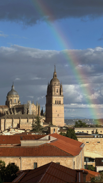 La imagen muestra un paisaje urbano con tejados de tejas rojas en primer plano y una catedral de piedra al fondo, dominada por una torre campanario y una cúpula. Un arcoíris surge del horizonte, cruzando el cielo parcialmente nublado con zonas de luz azul y nubes grises, creando un contraste llamativo sobre la arquitectura histórica.