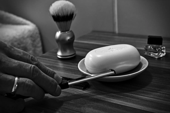Eine Hand hält ein Rasiermesser, das noch auf einer Seifenschale liegt. In der Schale liegt Rasierseife, noch trocken und neu. Links daneben ein Rasierpinsel. Hinter der Seifenschale steht ein kleiner Parfümflakon.