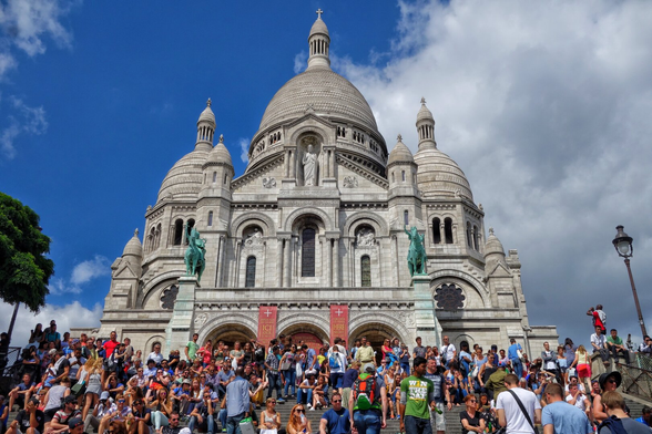 The image shows the Sacré-Cœur Basilica in Paris, a grand white-domed church with intricate architectural details, perched atop the Montmartre hill. Its large central dome is flanked by smaller domes and ornate towers. In the foreground, a bustling crowd gathers on the wide steps leading to the basilica, with people sitting, standing, and taking photos. The sky above is a mix of bright blue and dramatic clouds, adding contrast to the scene. Green statues of historical figures stand near the entrance, and a classic Parisian streetlamp is visible to the right.
