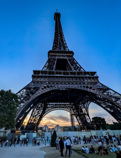 A low angle shot looking up at the Eiffel Tower at dusk. In the background, a blue sky with an orange sunset glowing through the base of the tower, while tourists gather in the foreground.