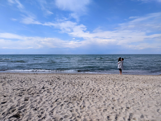 A serene view of the Baltic Sea at Dierhagen, with a wide, sandy beach in the foreground. The sand is light-coloured and textured with footprints. A woman stands near the water’s edge, holding a child, both gazing out at the calm, blue-grey sea, which stretches to the horizon under a bright sky with wispy clouds.