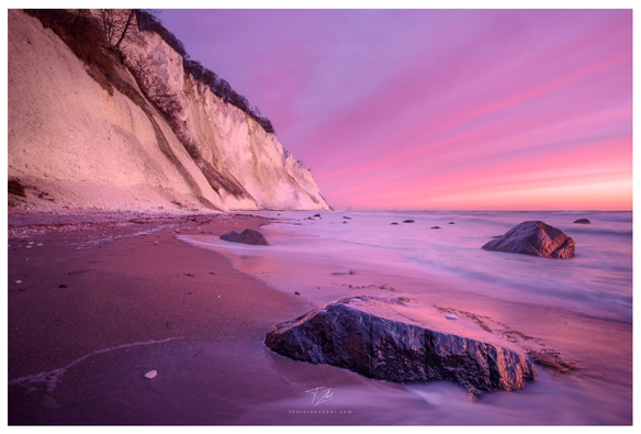 The photo was taken from the beach. In the background, you can see the cliffs of a rugged coastline, while the foreground is dominated by large boulders lapped by the waves. The sun, just about to rise, bathes the scene in shades of orange and magenta.