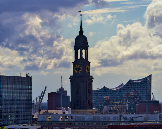Blick über Hamburg - vorn zentral der Turm des Hamburger Michels, dahinter ragt das markante Dach der Elbphiöharmonie in den locker bewölkten Himmel hinein. 
Die Turmuhr des Michels zeigt, dass es gerade zwölf Uhr ist. 
Rund um diese beiden sehr bekannten Gebäude sind weitere, weit weniger bekannte und auch Hafenkräne zu sehen. 