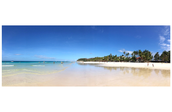 Panorama shot of a serene seaside vista. A calm ocean sweeps in from the left towards a beach of white sand with an extremely gentle slope. Tiny, distant beachgoers stroll along the sand, or wade in the water that remains ankle-deep even far out from shore. Palm trees stand over houses and low-rise structures at the horizon, their images reflected in the shallow water. A distant boat floats off to the left. Above is a clear blue sky with a few puffs of white clouds. The scene is in vivid hues of blue, teal, and green.