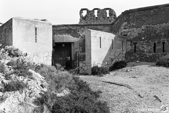 The entrance to Castillo de San Ramón with a wooden door, surrounded by rocky terrain and sparse vegetation. The remnants of an old fortification rise above, featuring broken walls against a stark sky. The image is captured in black and white, emphasizing its historical significance.
