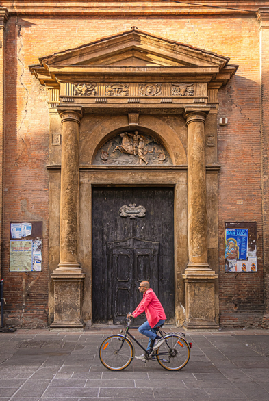 This is a photograph of the side door at San Martino Maggiore in Bologna. In the foreground a man wearing a bright pink shirt and blue jeans is riding a bicycle in front of a weathered, ornate brick building (the church). He is passing in front of the large, dark wooden door. The door is framed by two tall, cylindrical columns and a decorative pediment with relief carvings - work by Francesco Manzini. There are two posters on the brick walls on either side of the door. The street is paved with grey stone.