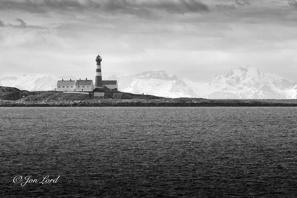This is a black and white photo in landscape format of the Landegode Lighthouse with a range of snow covered mountains in the background. Vestfjorden, Norway (2023).

The lower half of the image is covered with an expanse of dark, ink black, sea water, with small wavelets disturbing the surface. A faux horizon is created where the sea touches a narrow strip of land at the halfway up level. The land is a small island that is flat on the right half of the photo, only rising above the water by a few metres. From the centre of the image the rocky terrain rises slightly, perhaps four metres above the sea. To the left of centre is a classic looking lighthouse: tall, cylindrical, tapered with increasing height and with light and dark horizontal strips circling the tower. To the left of the lighthouse are two white, single floor buildings with grey pitched roofs, probably houses for the keepers in the past. Behind is a similar sized building without windows, so possibly a storehouse. In front are two small outbuildings. In the distant background, about 50km away, is a range of snow covered mountains stretching from the left to the right of the image. 

The lighthouse, located 18km north of Bodø, in Norway's Arctic north, was built in 1902, is made from cast iron and is 29 metres tall. The body of water beyond the light is the Vestfjorden. The mountain range, often called the 'Lofoten Wall', is on the southern part of the Lofoten archipelago.
