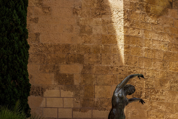 Español: muro de piedra arenisca iluminado por una franja vertical de luz solar que lo divide en zonas de sombra y resplandor. En la esquina inferior derecha, una escultura de bronce de figura humana con el cuerpo arqueado y el brazo alzado, como en movimiento.

English: sandstone wall divided by a sharp vertical beam of sunlight casting contrasting zones of shadow and golden light. In the lower right corner, a bronze sculpture of a human figure with arched body and raised arm, caught in motion.