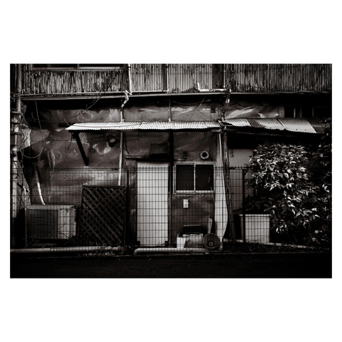 A black-and-white view of a weathered building facade with corrugated metal roofing, patched walls, exposed wiring, and uneven wooden supports. A chain-link fence runs across the foreground, partially blocking air conditioning units, small windows, and stacked utility fixtures. On the right side, a dense shrub grows beside the structure, adding texture against the layered metal and wood surfaces.