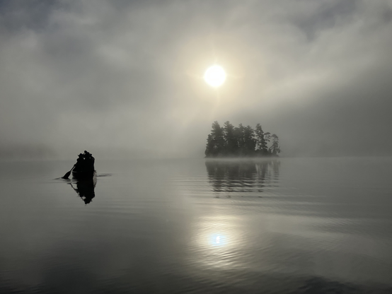 Looking towards the sun across a calm lake with the silhouette of an island in the distance on  the right, and a the silhouette of a canoe with two paddlers paddling into the distance on the left. The sun is behind rising fog.