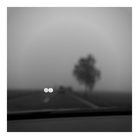 A black-and-white photograph taken through a rain-covered wind screen. The image shows a blurred view of a tree and a road with two bright headlights visible in the distance.