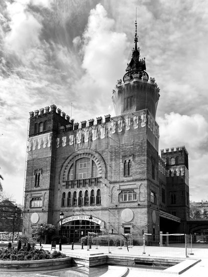 This black and white photo shows a Castell in Barcelona. It is a wonderful fortress. A fountain can be seen in the foreground. The weather is spring-like, the sun is shining and it is cloudy. The clouds above the Castell draw a beautiful pattern in the sky.