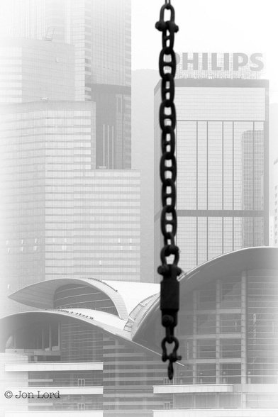 This is an abstract black and white photo in portrait format of a distant high-rise cityscape with a heavy duty chain suspended in the foreground. Wan Chai, Hong Kong (2012).

From a point about one third from the right edge of the photo is a black, heavy chain that extends downward to a point just above the base of the image. The chain is slightly blurred out of focus but the multiple 'stud links' (the type of heavy chain link used to anchor ships) are clearly visible. The chain is hanging from the boom of a derrick barge that's out of view above. In the background, 1km away and visible through a thin haze, is a densely spaced cityscape made up of multiple high-rise, glass fronted buildings. One of the skyscrapers on the right has the words 'HILIPS' in large bold letters, the chain is obscuring the first letter 'P'. In front of the high-rises is a futuristic, low-rise building with an uneven , faceted front with three vast wave or wing like white roofs.

The cityscape are the many high-rise blocks that line the Victoria Harbour waterfront of Wan Chai on Hong Kong island. The futuristic building in front is the Hong Kong Convention and Exhibition Centre.


