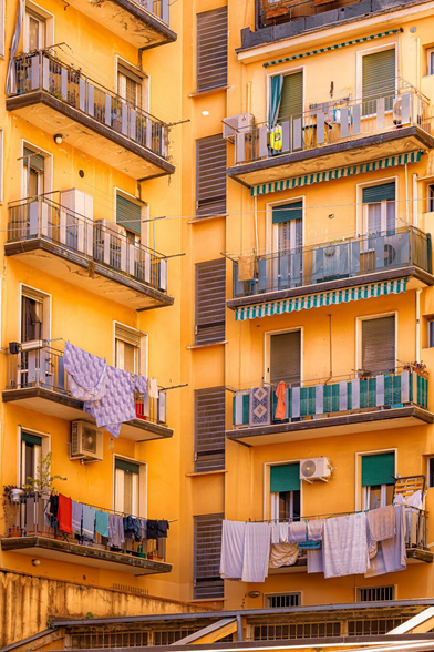 This is a photograph of a sunlit, yellow-orange, multi-storey apartment building with six balconies (which are visible). Each balcony has something different on it. Some have laundry, including striped and patterned clothes, hanging on rails and others have bits and bobs and plants. The buildings have green blinds and grey metal railings.