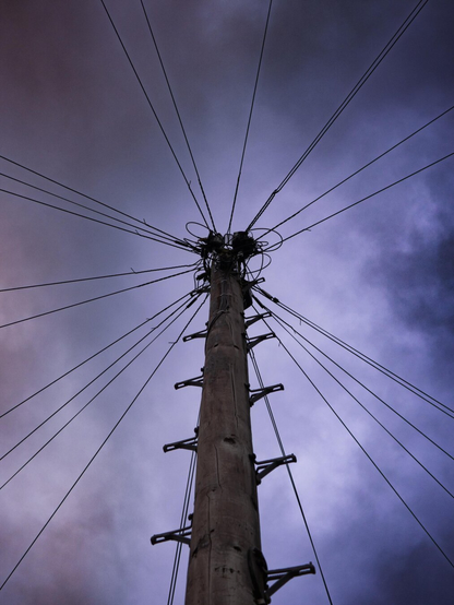 A weathered wooden telegraph pole with footholds shot from a low angle, showing numerous black telecommunication wires radiating outward from its top against a dramatic, cloudy sky in purple hues.