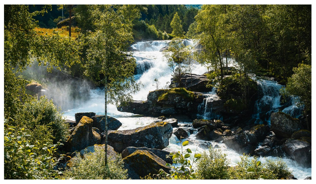 Wasser fließt in mehreren kleineren und größeren Kaskaden zwischen großen Steinen durch eine üppig grüne Landschaft.