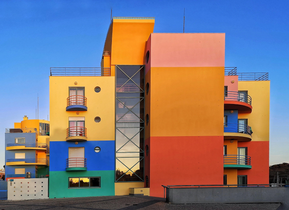 Das Foto zeigt ein buntes, mehrstöckiges, modernes Gebäude.
Die Fassade ist in verschiedene horizontale Farbquader und -quadrate unterteilt, darunter Gelb, Orange, Rosa, Blau und Grün. Ein markantes Merkmal ist der gläserne Treppenhausschacht in der Mitte des Gebäudes. Darin spiegelt sich der Sonnenuntergang. Die Fenster sind teilweise rund oder mit kleinen Balkonen versehen. Der Himmel im Hintergrund ist klar und blau.
