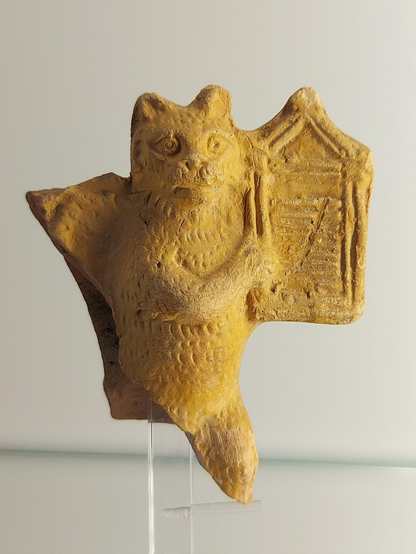 Fragment of a yellowish-brown clay figurine depicting a cat playing a harp. The cat is upright, with detailed facial features and textured fur, holding the harp to its side. The surface shows signs of wear and age, and the piece is displayed on a clear stand.