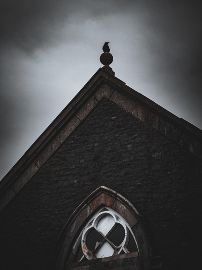 A lone crow sits atop the finial on the peak of a weathered, abandoned stone chapel. It is centered under a dark and moody overcast sky.