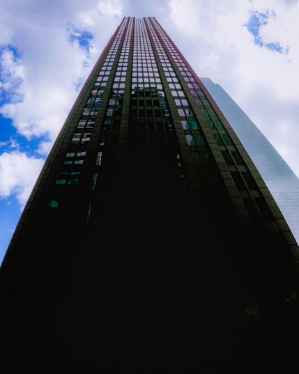 Low-angle photo of a dark skyscraper stretching into a cloudy blue sky, with grainy texture and reflective purple-tinted windows that give the image a nostalgic early-2000s digital look.