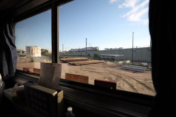 View through the window of a roomette on the westbound Amtrak California Zephyr headed out of Denver. A dirt lot stocked with construction materials is bordered behind by a raised railway. On that railway is another train, one of the A trains to Denver International Airport.