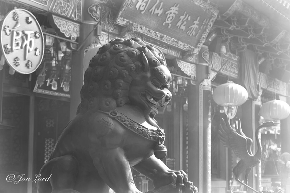 This is a black and white photo in landscape format of the interior courtyard of a Taoist temple with a fierce lion in the foreground and an ornate oriental facade, partially obscured by a haze of incense smoke. Wong Tai Sin Temple, Hong Kong (2017)

In the foreground and centre of the photo is a large statue of a lion-like mythical creature, about the size of an adult lion, facing to the right, viewed obliquely with the front and right side in view. The creature has its large mane arranged in curls, its mouth is open as if snarling and revealing teeth and a tongue. In the near background, a few metres behind, is the ornate front of the temple, again viewed obliquely, with the left portion being closest to the camera and the facade becoming more distant towards the right. The facade overhangs a covered entrance and walkway. It is highly ornate with round columns and square pillars supporting a large carved, wooded lintel. In the upper left corner is a round plate hanging from the lintel with one large Chinese character in the centre and 8 smaller characters evenly spaced around the edge. Over the entrance is a large rectangular sign with small delicate embellishments around the 4 edges and 5 large Chinese characters within. Two large Chinese paper lanterns are hanging either side of the entrance with a life sized statue of stork or similar bird, wings outspread and long neck extended upwards. All of this is viewed through a thin, but noticeable haze of incense smoke.