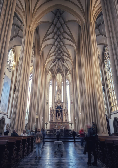 Interior of Saint James' Church (Kostel sv. Jakuba) in Brno, featuring towering Gothic arches, intricate ribbed vaulting, tall stained-glass windows, and a central ornate altar, with visitors scattered throughout the space.