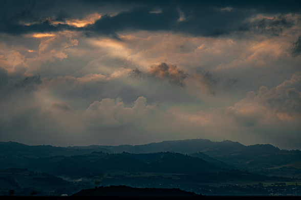 The photo is of a dramatic sky filled with a mix of dark and some warm, golden light breaking through. The sky fills up the top two-thirds of the frame, more or less. 
Below the sky, there are rolling hills and mountains which are silhouetted. 
The description is short because I am just back and tired 😂