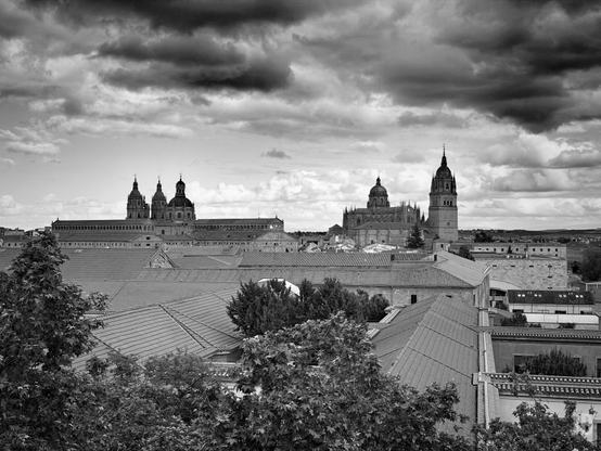 La imagen muestra una vista panorámica en blanco y negro de una ciudad histórica con tejados de teja y árboles en primer plano. Al fondo destacan varias torres y cúpulas monumentales, entre ellas una gran catedral, bajo un cielo dramático con nubes oscuras y densas que crean un fuerte contraste.