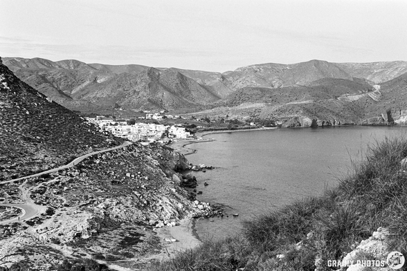 A black and white landscape featuring rugged mountains and a serene coastline. A winding road leads to Las Negras by the water, surrounded by cliffs and greenery, showcasing the natural beauty of the area.