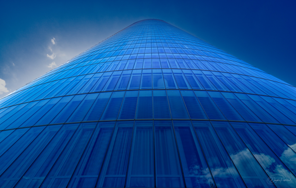 A view from the bottom of a modern tall glass skyscraper with a facade of blue tinted glass reaches far into the sky blending with the deep blue of the sky itself.  White puffy clouds are reflected in some of the panes.  The building appears to glow in the sunlight.
