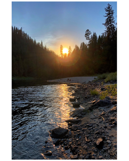 The Clearwater River at sunset. The water ripples alongside a stony riverbank in the foreground that recedes to a sandy shore in the distance, on which two tiny people are relaxing. A shadowy mass of tall conifer trees rises behind them. The line of treetops dips in a “V” shape with the golden sun nestled at the bottom of the V, casting a column of glittering light on the water. The sky above is a deepening blue with wispy clouds.