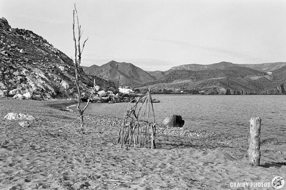 A black and white photograph of a serene beach scene featuring a drifteood sculptures, including a structure resembling a small shelter, sandy shore, scattered rocks, and distant mountains under a clear sky.