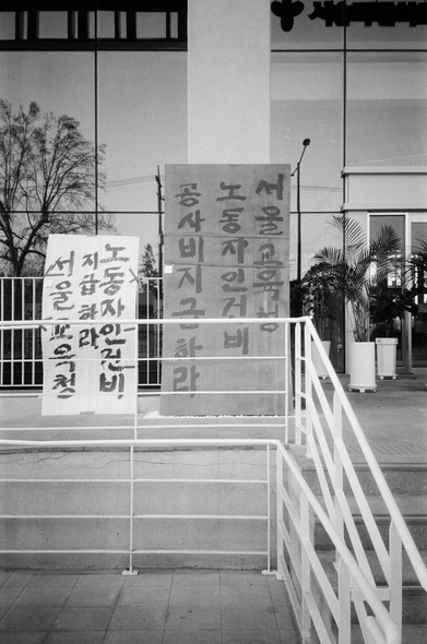 서울시교육청 입구 옆에 붙은 거대한 판자 2개에 세로쓰기·손글씨로 메시지가 써있다.
“서울교육청 노동자 인건비, 공사비 지급하라”
“노동자 인건비 지급하라 서울교육청”