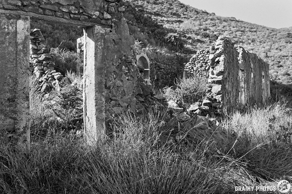 Black and white image of abandoned stone ruins partially covered by tall grass, situated on a hillside. The remnants showcase walls and doorways, evoking a sense of decay and natural reclamation in a serene landscape.