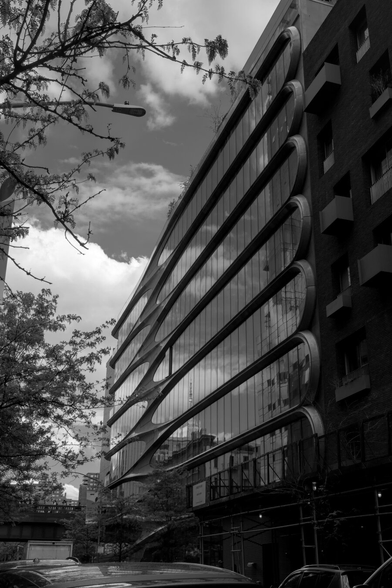 High contrast black and white photo showing a mid-rise building with streamlined patterns around its shiny glass walls, reflecting a partially cloudy sky