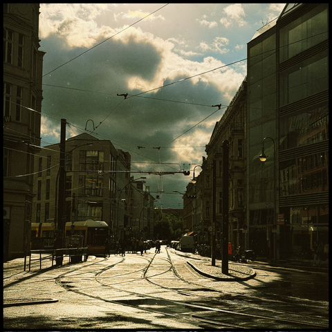 Regennasse Straße mit Straßenbahnschienen im Gegenlicht, links und rechts Hausfassaden und eine schwere Wolke am Himmel