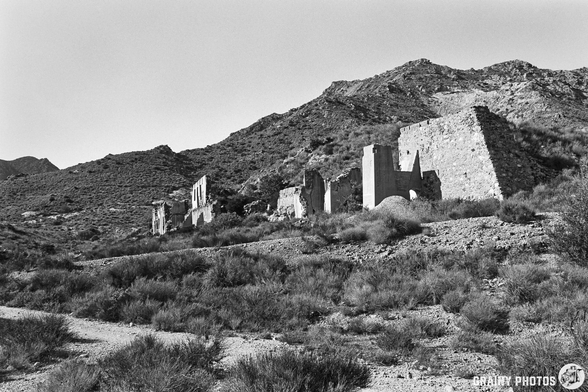 A black-and-white photograph of crumbling ruins on a hillside, surrounded by sparse vegetation and mountainous terrain under a clear sky, evoking a sense of abandonment and the passage of time.