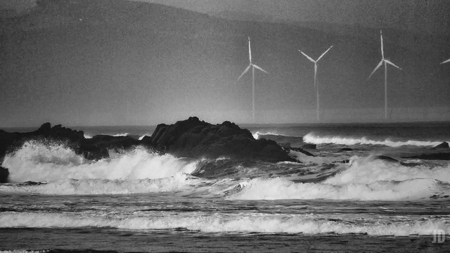 La imagen muestra un paisaje costero en blanco y negro, con olas rompiendo contra rocas oscuras en primer plano. Al fondo, sobre el horizonte marino, se distinguen varios aerogeneradores, parcialmente difuminados por la distancia o la neblina. La escena transmite una sensación de fuerza natural combinada con elementos de energía renovable.