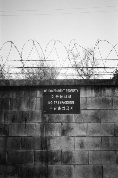 미군기지 담장. 큼직한 콘크리트 블럭으로 쌓여있고 위쪽에는 철조망이 설치되어 있다. 아래쪽에 팻말이 붙어있다. “US GOVERNMENT PROPERTY 미군용시설 NO TRESPASSING 무단출입금지”