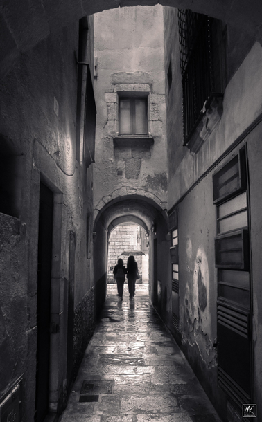 Monochrome photo looking down a covered arched stone passageway with two people at the far end about to step out into a street, one is holding an open umbrella. 