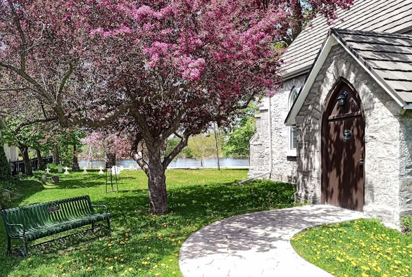A crabapple tree covered in pink blossoms stands beside a small church. Yellow dandelions can be seen in the grass and a bench sits on the left side. A river can be seen in the distance. 