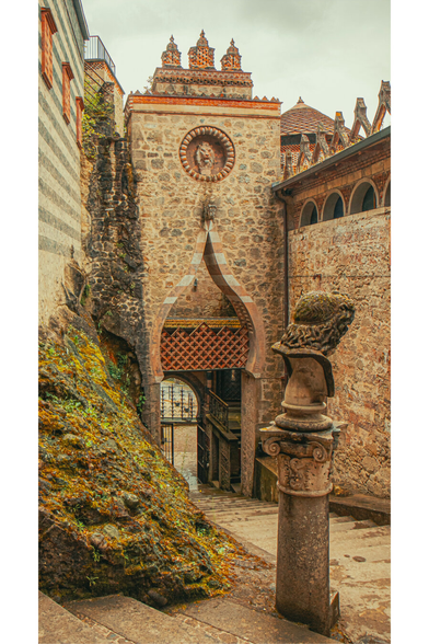 A phototograph of the entrance into La Rocchetta Mattei. There is what appears to be a medieval stone archway (actually it was built in the 19th century!) with a circular sun emblem with a lion statue above it -set in a narrow, cobblestone alley like entrance. The arch has a Moorish-style pointed top and an arched metal lattice part. To the right there is a weathered stone column with an ornate, carved head. The background has moss-covered walls and red-tiled rooftops with intricate architecture.